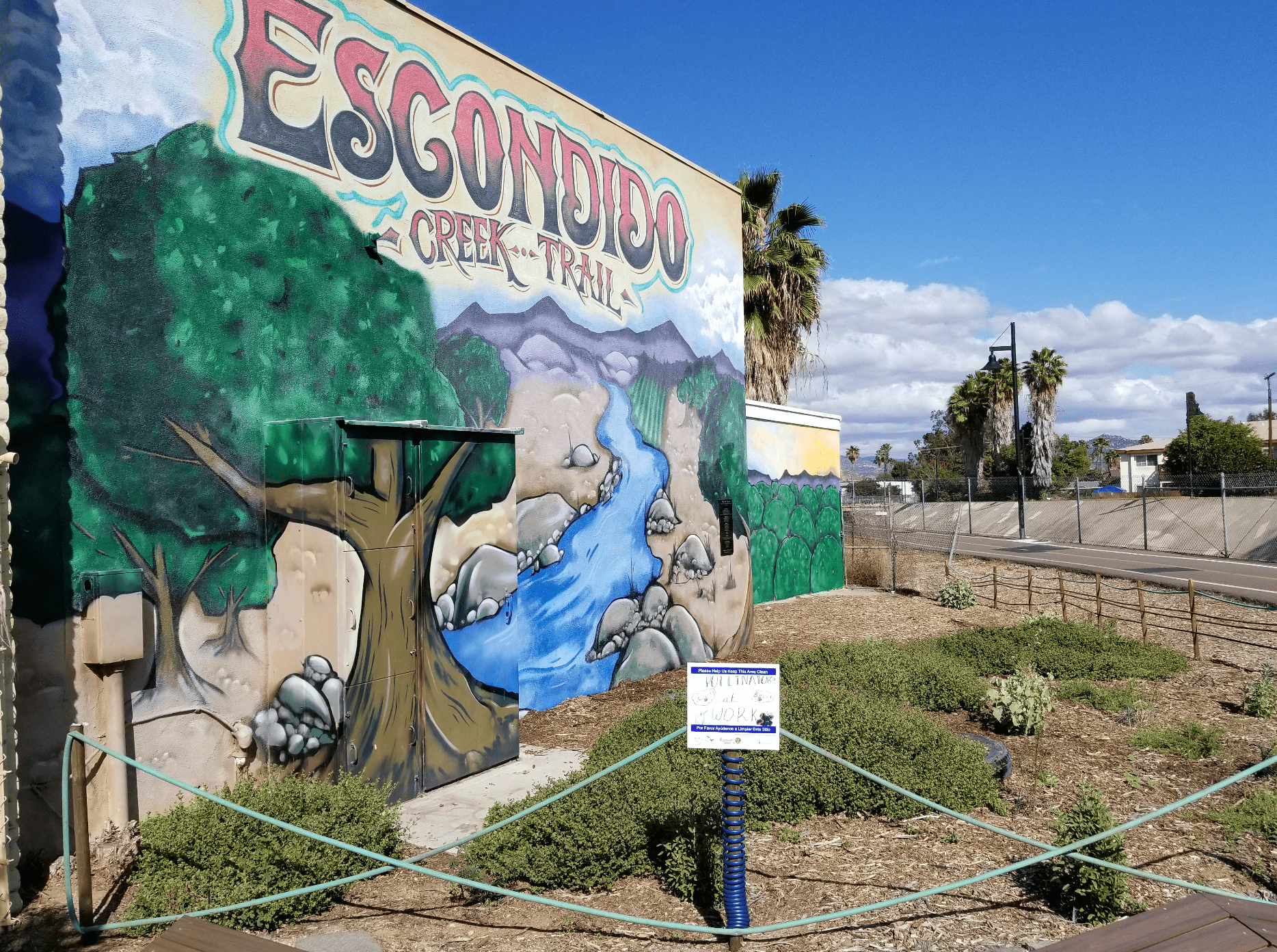 A pollinator garden in the foreground leads your eye to a mural on the side of a concrete block building showing a blue creek leading off into a mountain range in the distance. Large trees frame the river.