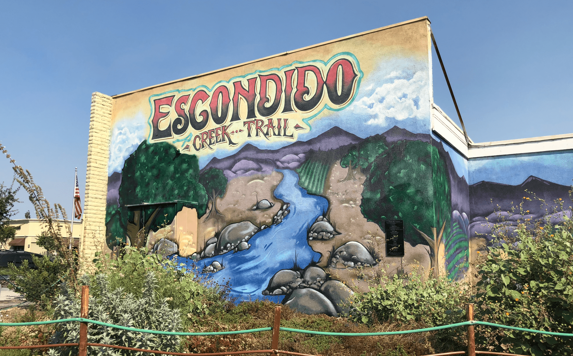 A large mural of a river, surrounded by trees and boulders leads up from the bottom of a concrete wall, disappearing into a mountain range.