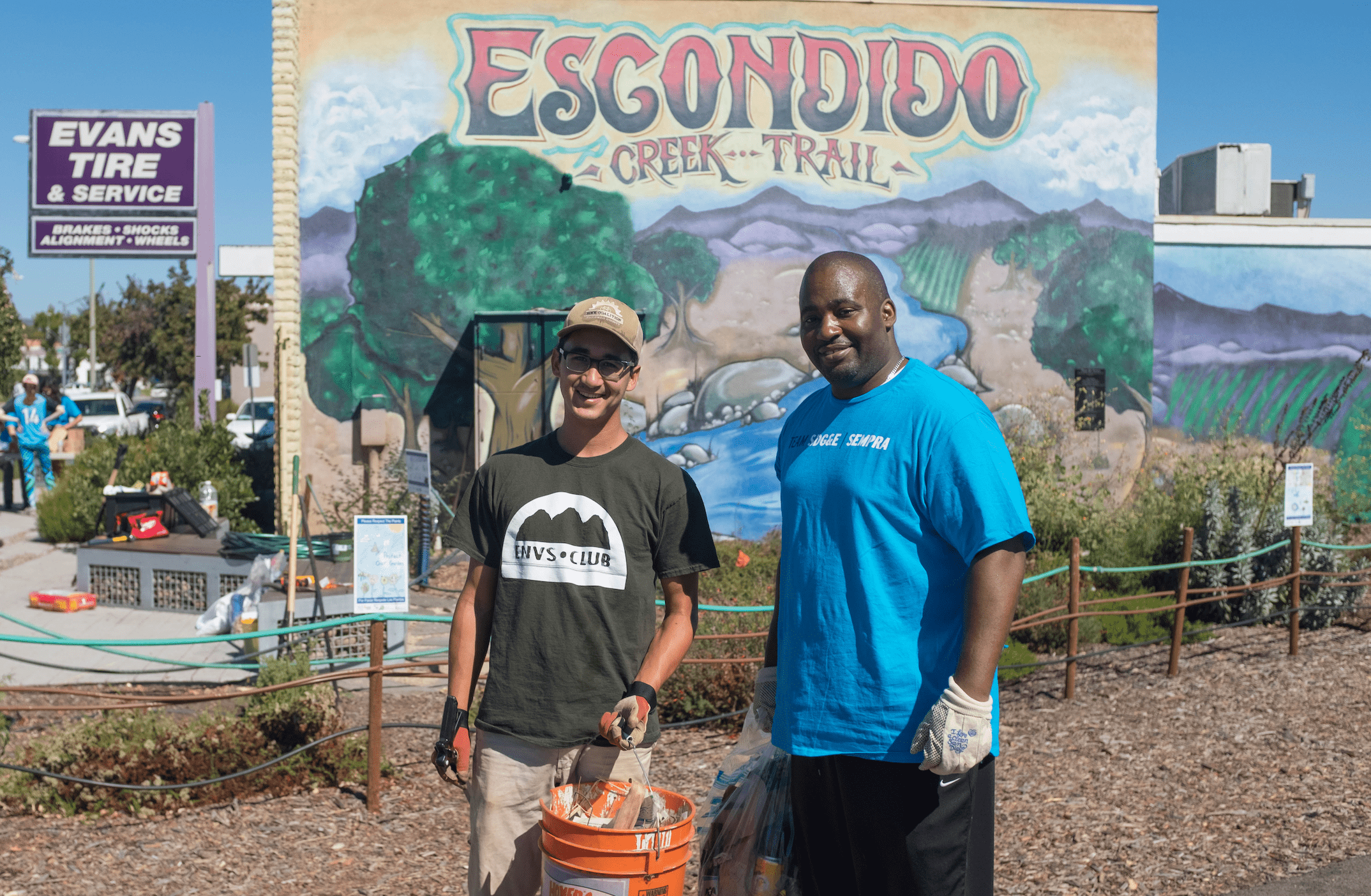Two volunteers holding orange trash buckets pose for the camera with smiles in front of a mural of a creek passing through a forest towards a mountain range.