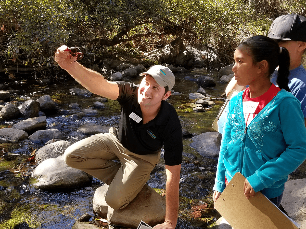 Escondido Creek Conservancy Education director crouches on a rock in the middle of a stream, while holding up a crustacean. Elementary school students look on.