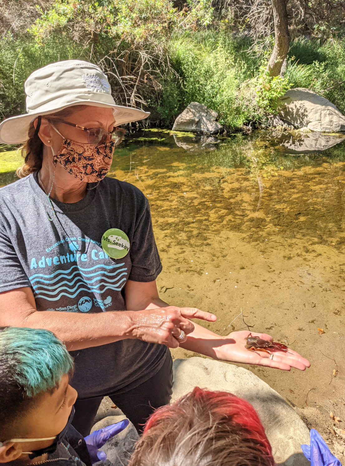 Lead educator holds a crayfish to be seen by students while standing in front of a creek.
