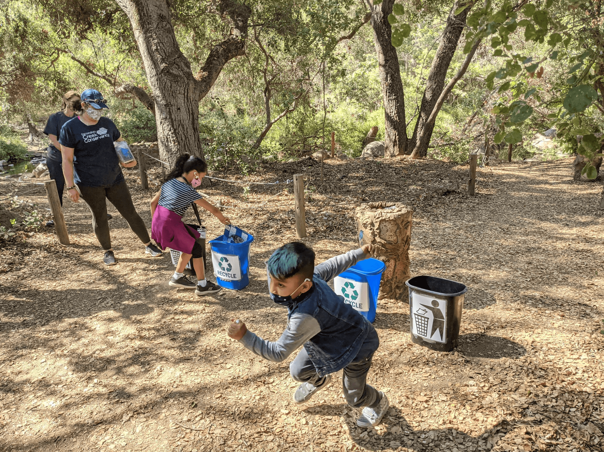Elementary age students running to pick up a piece of recycling on the ground under a canopy of trees. A row of recycle bins cuts through the center of the image. One student is running towards the camera, while another is dropping a piece of recycling in the buckets. Other students and camp counselors look on.