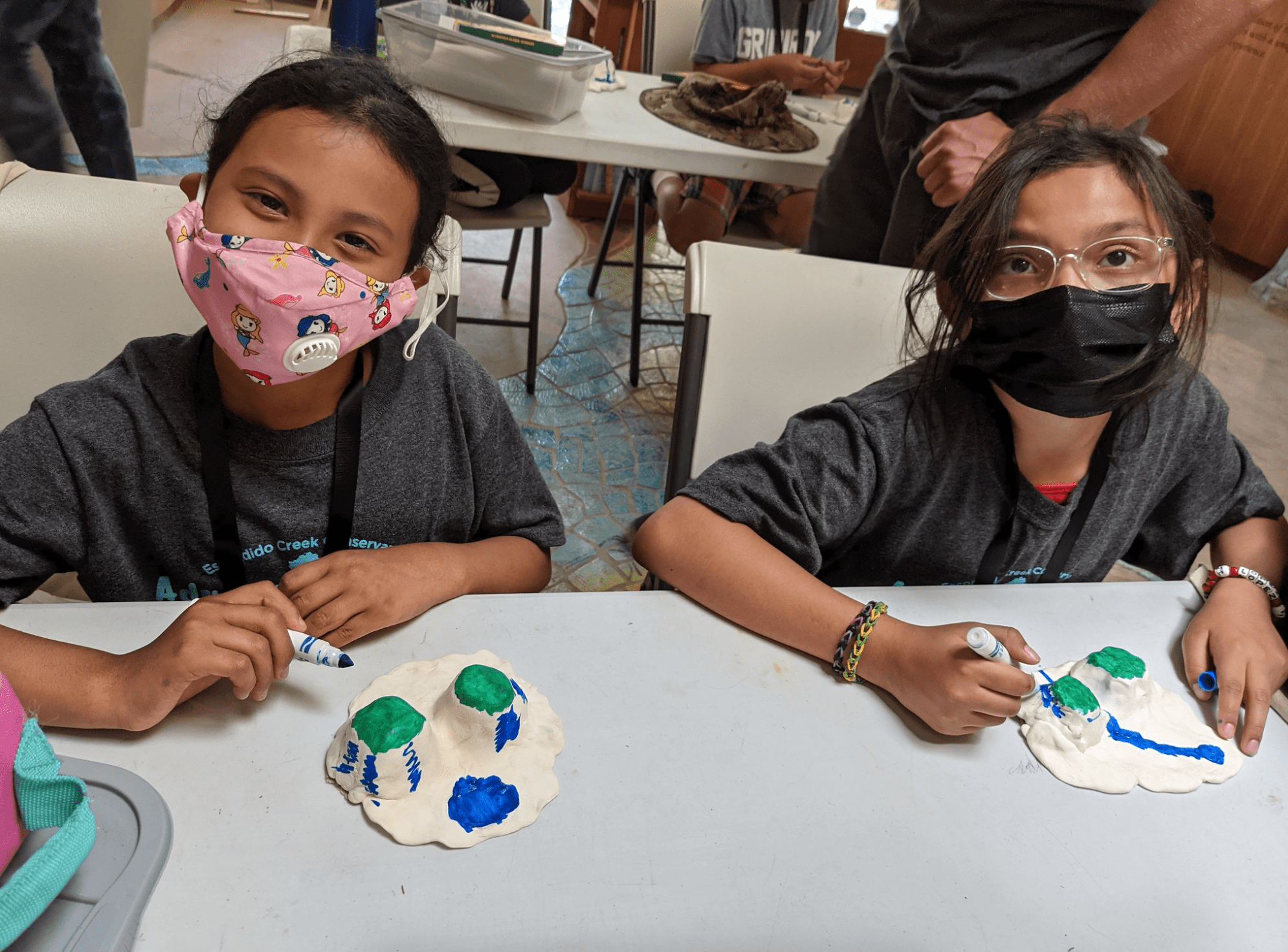 Two elementary age students paint clay and smile at the camera.