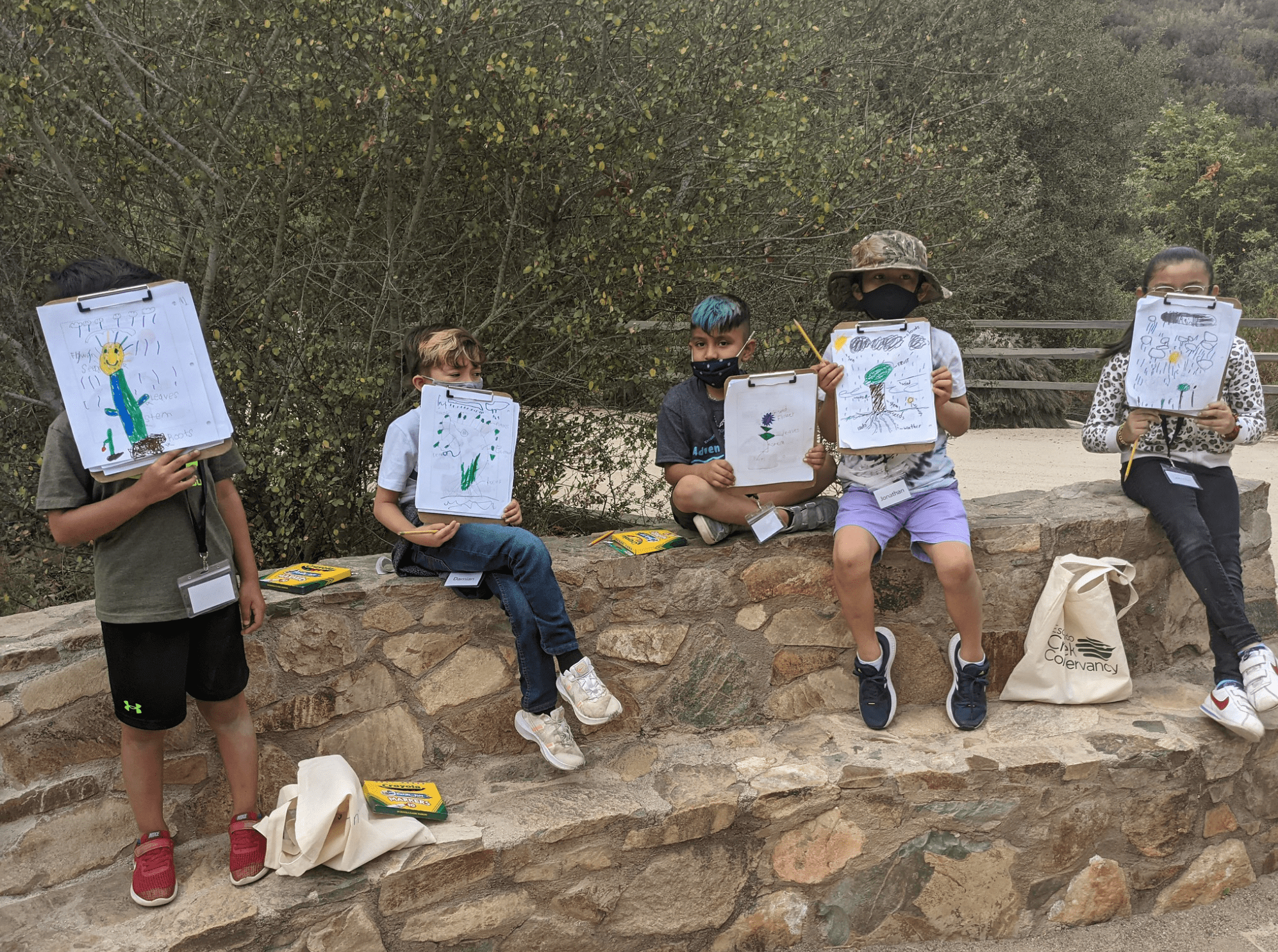 Students sit on a rock ledge holding up their drawings of nature.