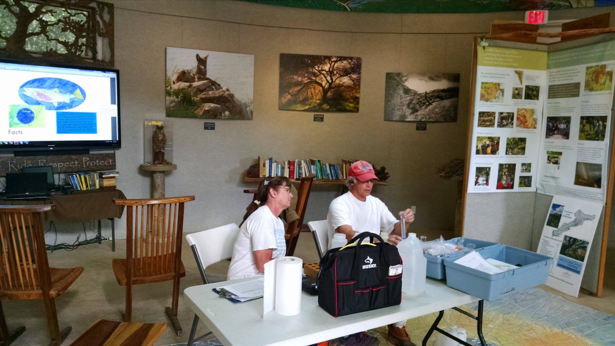 A man and a woman work with water quality testing tools at a card table in an environmental interpretive center.
