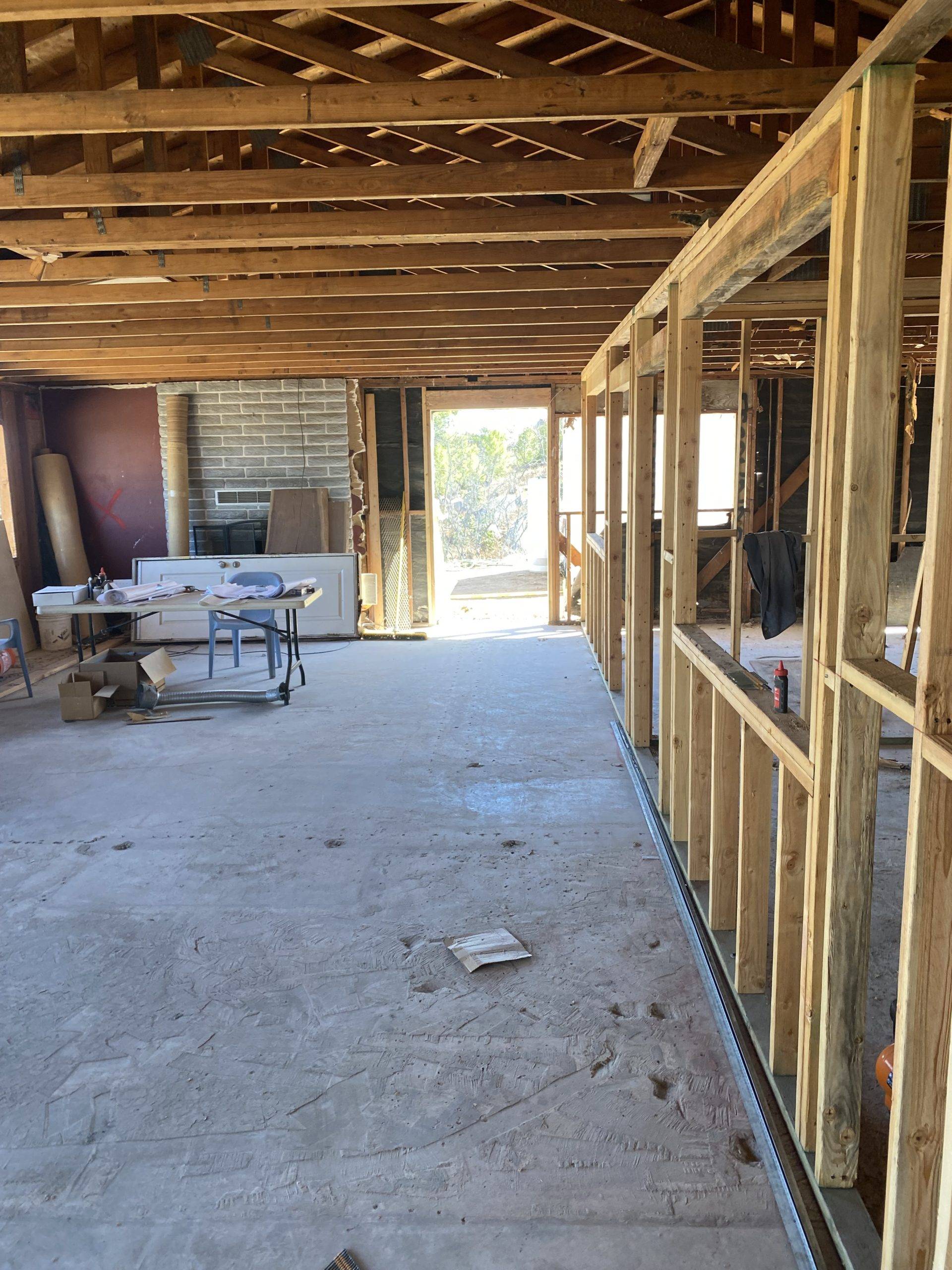 Interior of a ranch home under construction. All the walls are studs and the floor raw concrete. Photo is vertical with a stud wall on the right and the room cropped down the middle on the left. Open beams in the ceiling can be seen at the top.