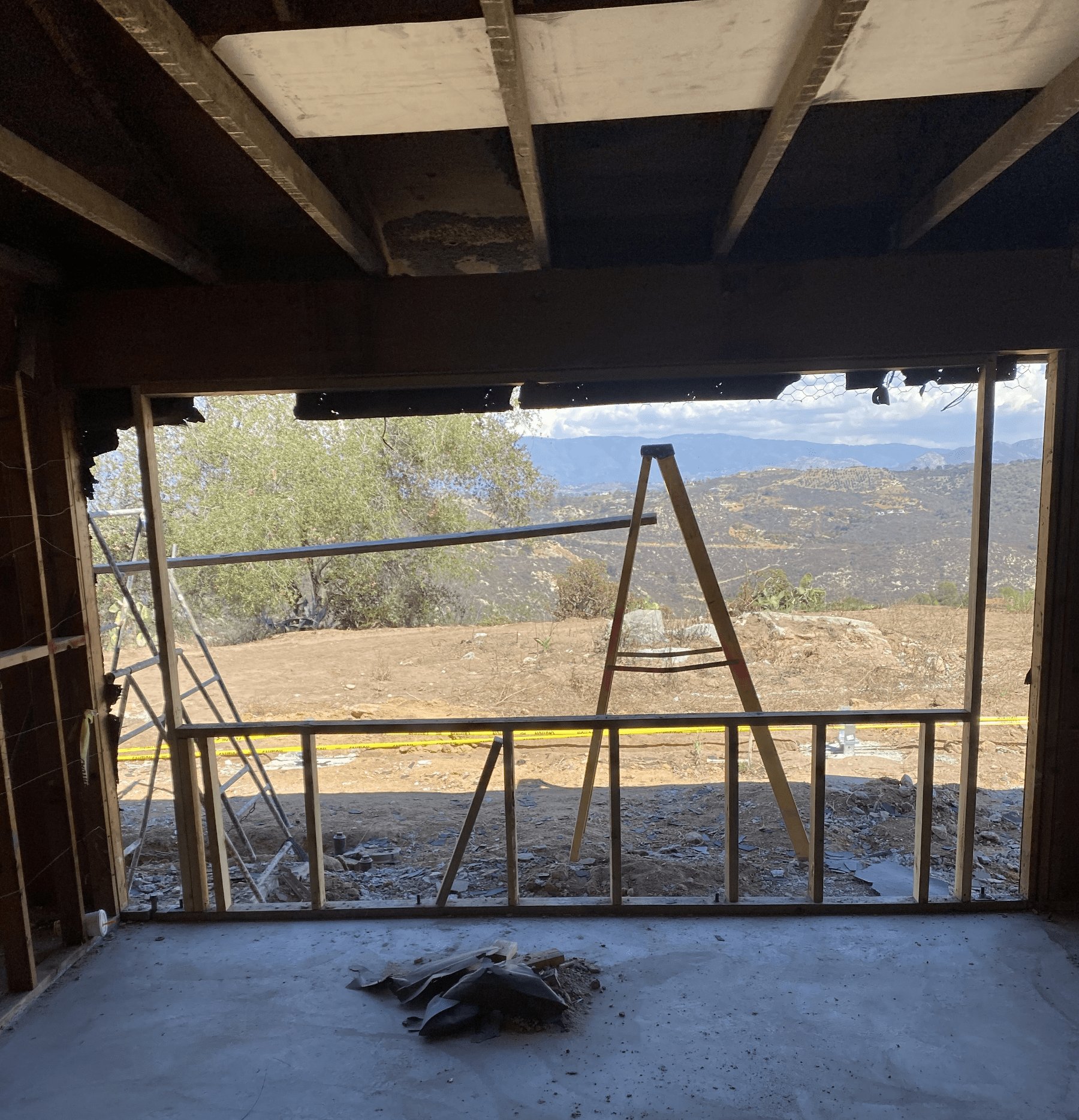 A photo from inside a construction site looking through a studded window frame at a mountainous view. An orange ladder and construction tools obscure the view slightly.