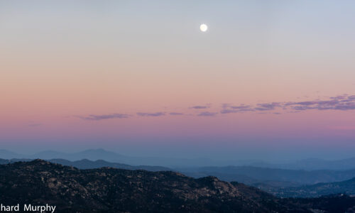 The sky just after sunset with streaks of blue, purple, red, orange, and the moon over head. The colors spread out over the low hills in east San Diego County.
