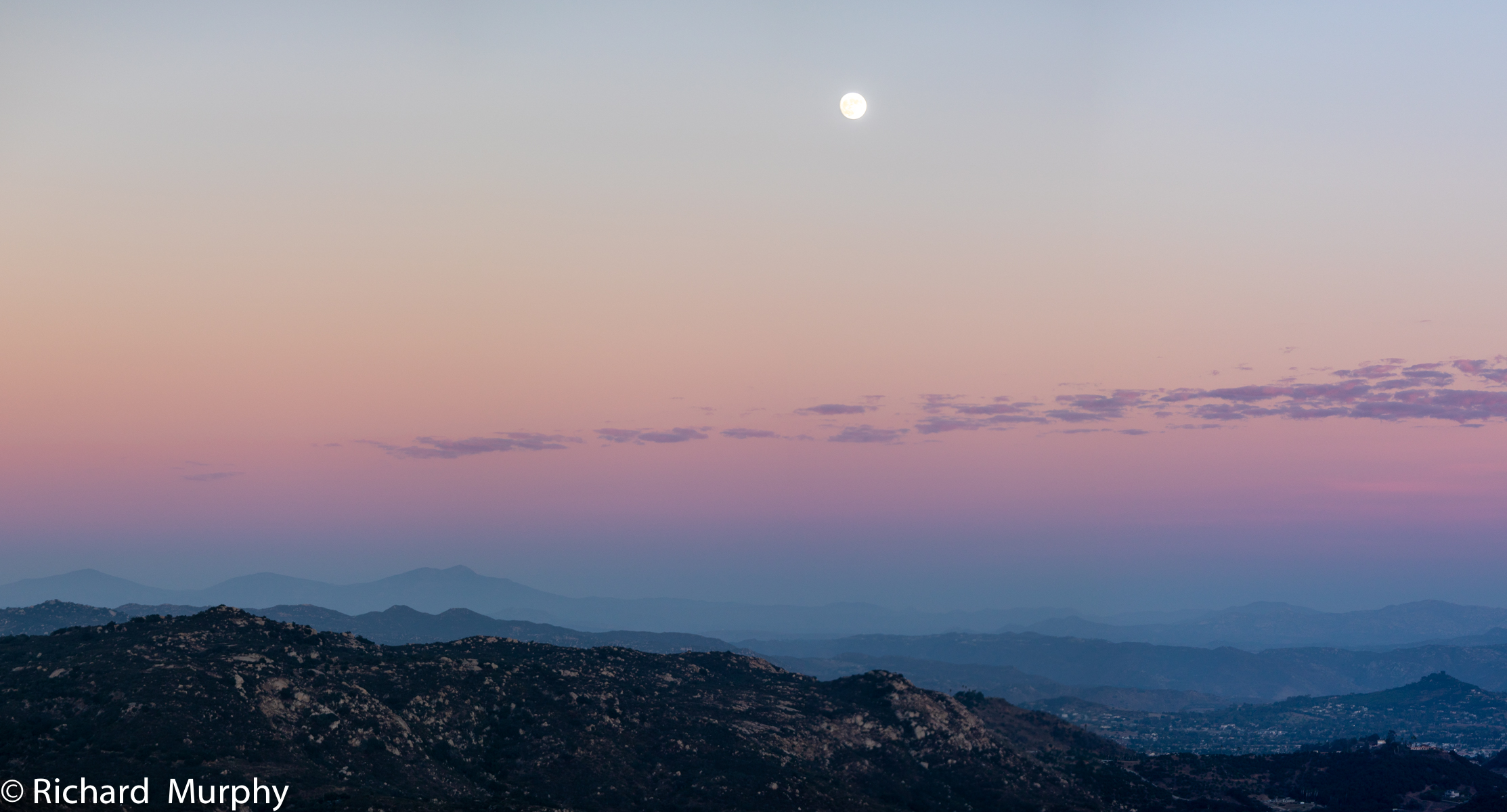 The sky just after sunset with streaks of blue, purple, red, orange, and the moon over head. The colors spread out over the low hills in east San Diego County.