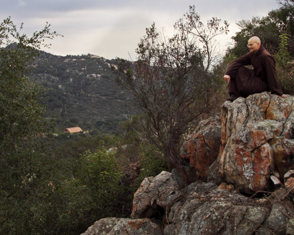 A monk sitting on a stone outcrop looking out over the wilderness.