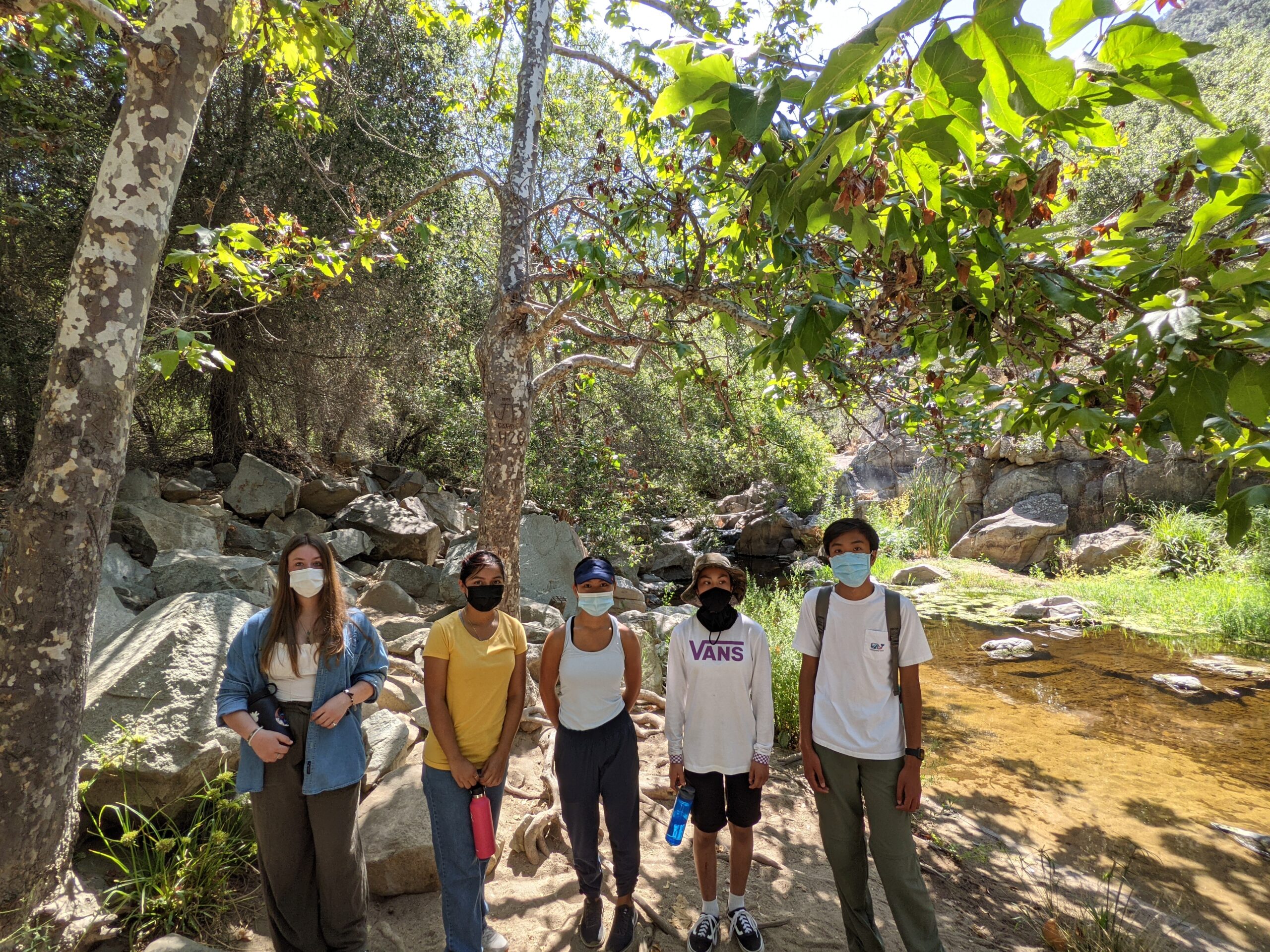 High school students pose in front of a creek in a shaded area of a regional park.