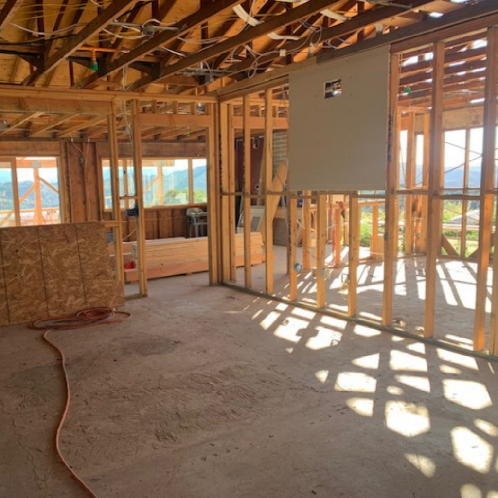 An empty room under construction with exposed studs, concrete floor, and views through the spaces where windows will be.