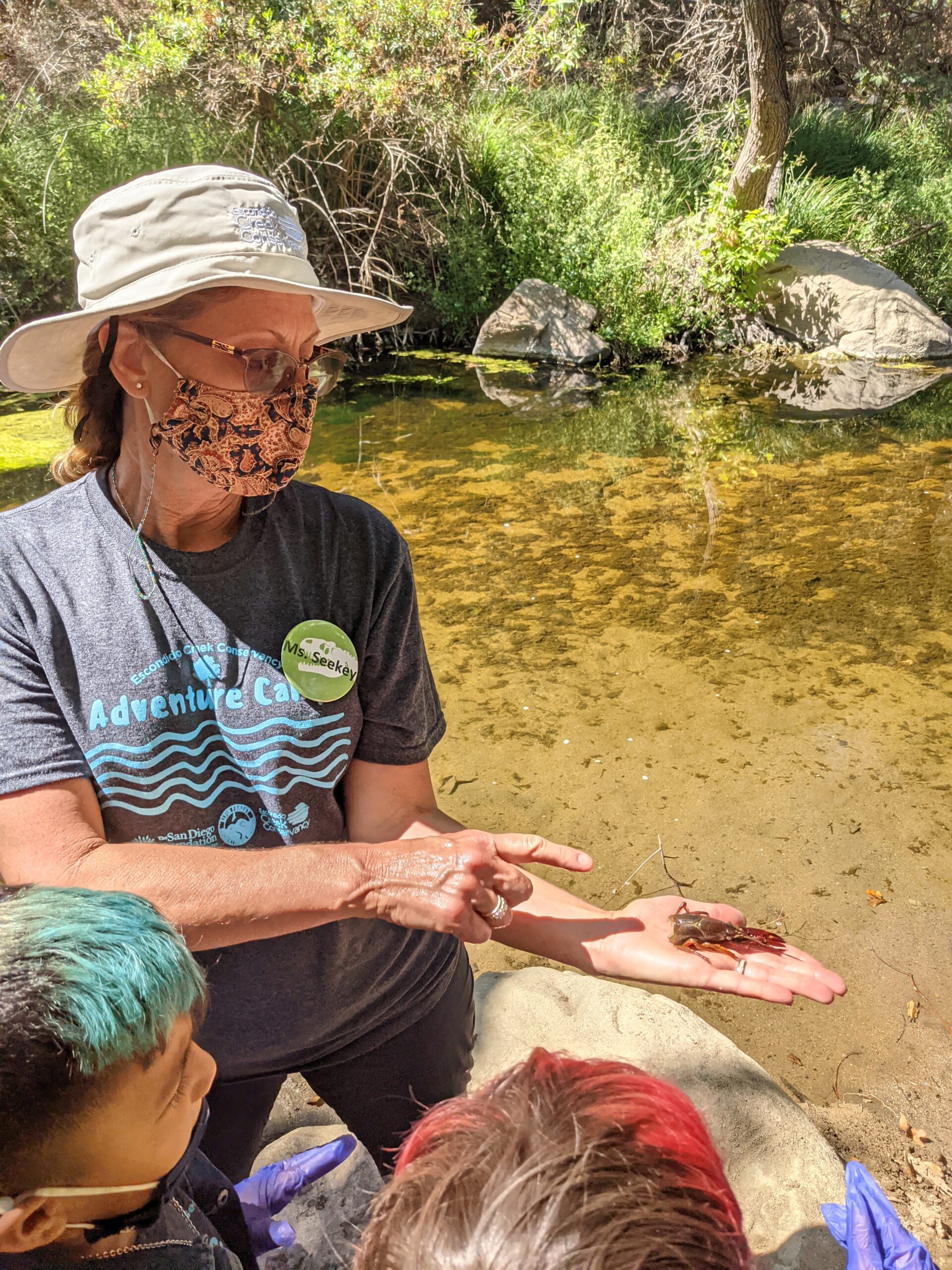 A teacher showing students a crawfish on her palm in front of a stream.