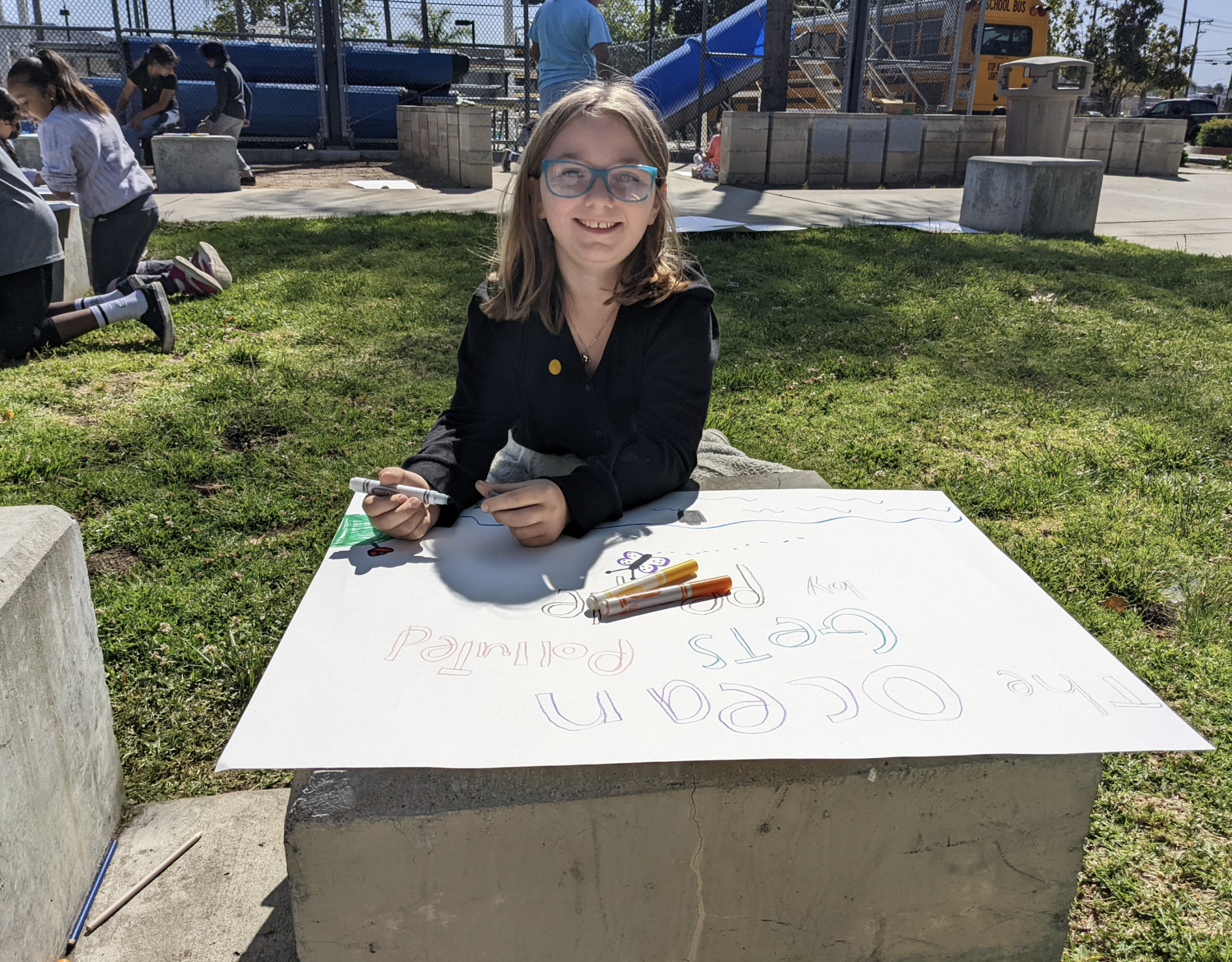 An elementary student draws a sign for Earth Day with marker on a big white board in the park.