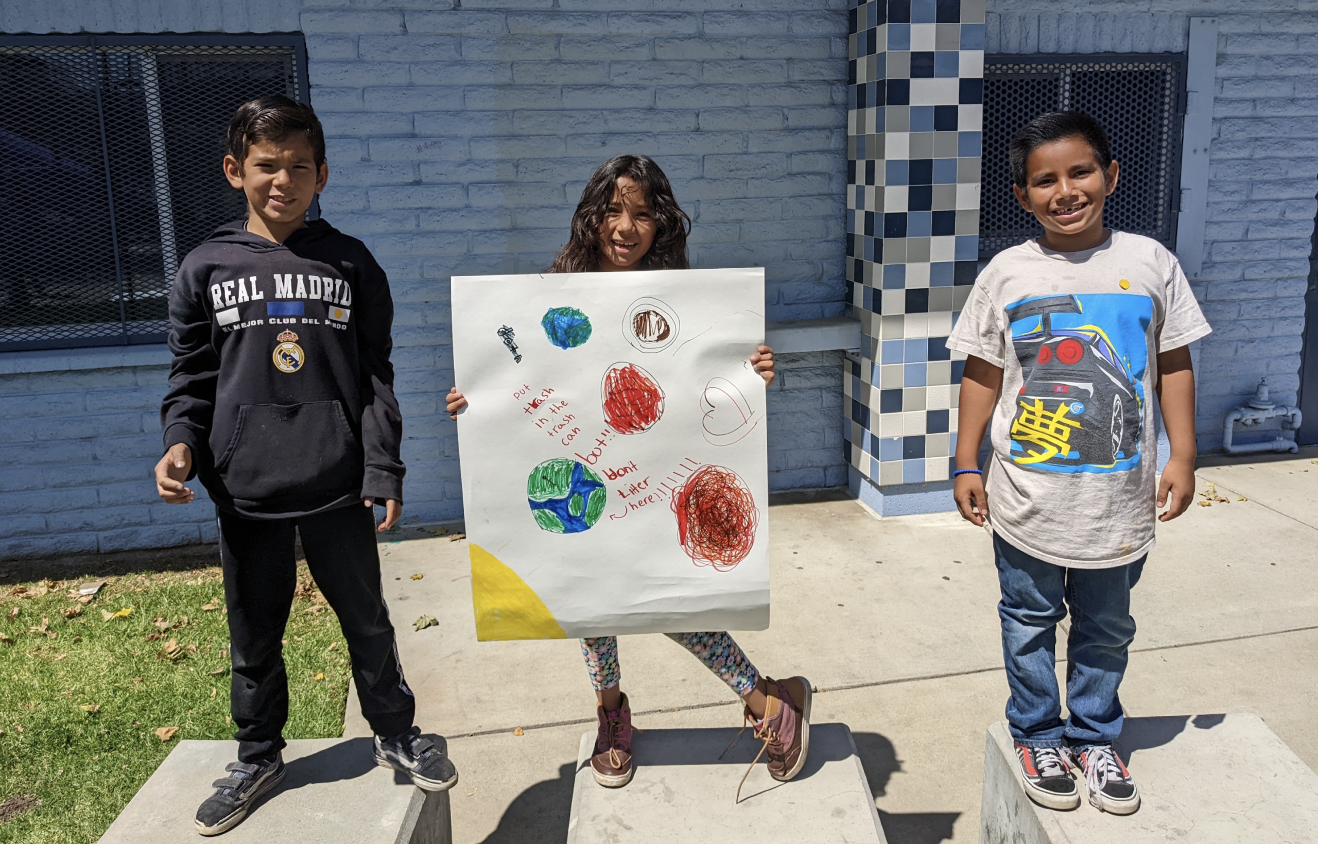Three elementary students stand side-by-side posing with their don't litter sign they designed for Earth Day.