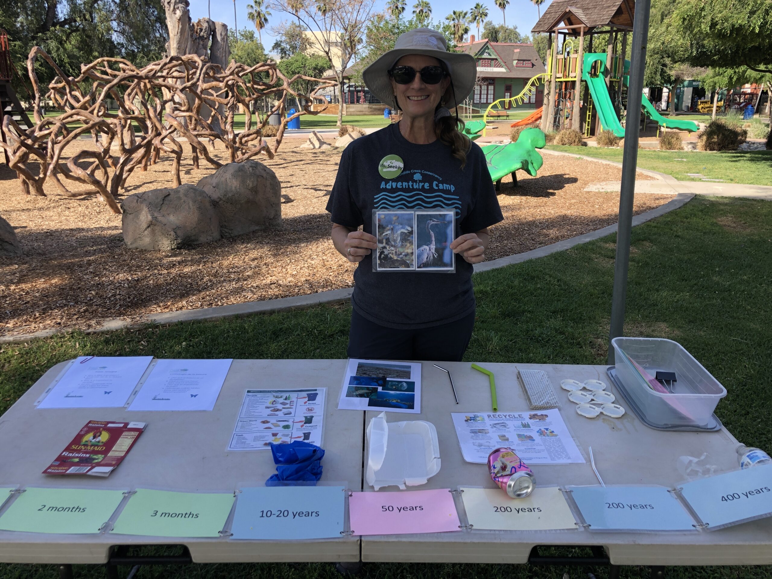 A woman holds up photos of native birds standing behind a table of environmental education pamphlets in front of a playground.