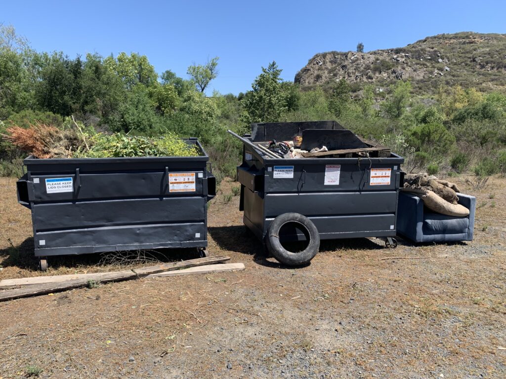 Overflowing black dumpster trashcans in front of scrub brush.