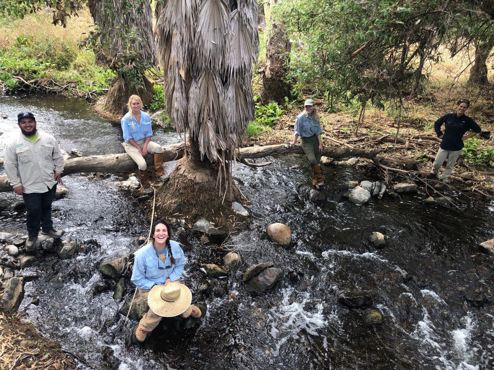 Members of a land conservation team look up at a the camera while standing and sitting on rocks spread across a running creek.